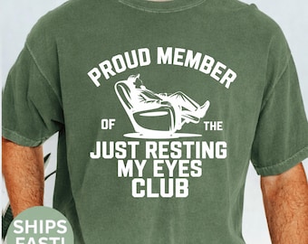 May include: Moss green t-shirt with white text and graphic. The text reads "PROUD MEMBER OF THE JUST RESTING MY EYES CLUB" with an illustration of a person relaxing in a chair. The shirt is a crew neck style.