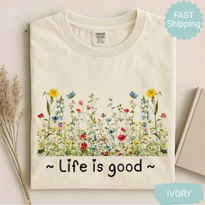 May include: An ivory t-shirt with a floral design across the chest. The design features wildflowers in yellow, red, blue, and pink. The text "~ Life is good ~" is printed below the flowers. The t-shirt is a Comfort Colors brand.