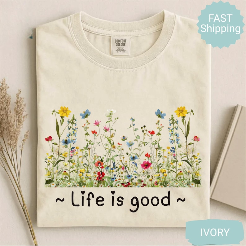 May include: An ivory t-shirt with a floral design across the chest. The design features wildflowers in yellow, red, blue, and pink. The text "~ Life is good ~" is printed below the flowers. The t-shirt is a Comfort Colors brand.