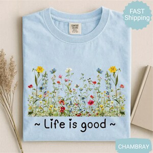 May include: Pale blue t-shirt with a vibrant floral design of wildflowers. The text "Life is good" is printed in black. The shirt is made of a soft material and is a comfortable fit.