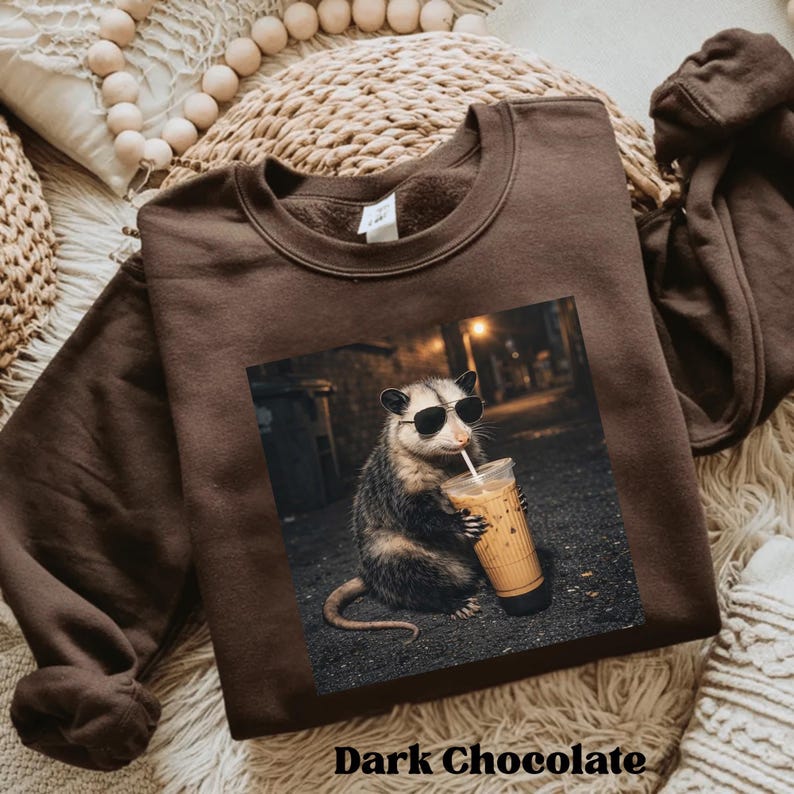 May include: Dark chocolate brown sweatshirt featuring a graphic of an opossum wearing sunglasses and drinking iced coffee. The image has a nighttime street scene background. The text "Dark Chocolate" is at the bottom.