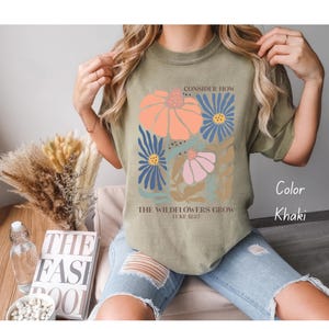 K&ouml;nnte beinhalten: Khaki-farbenes T-Shirt mit einem Blumenmuster und dem Text "Betrachte, wie die Wildblumen wachsen Lukas 12:27"
