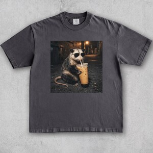 May include: Dark gray t-shirt featuring a graphic of an opossum wearing sunglasses, sipping an iced coffee. The opossum is in an alley setting. The shirt is a solid color with a crew neck and short sleeves.