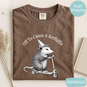 Könnte beinhalten: Espressofarbenes T-Shirt mit dem Text "Off To Cause A Kerfuffle". Die Grafik zeigt ein Opossum mit Hexenhut, das Roller fährt. Das Shirt ist aus Comfort Colors Material.