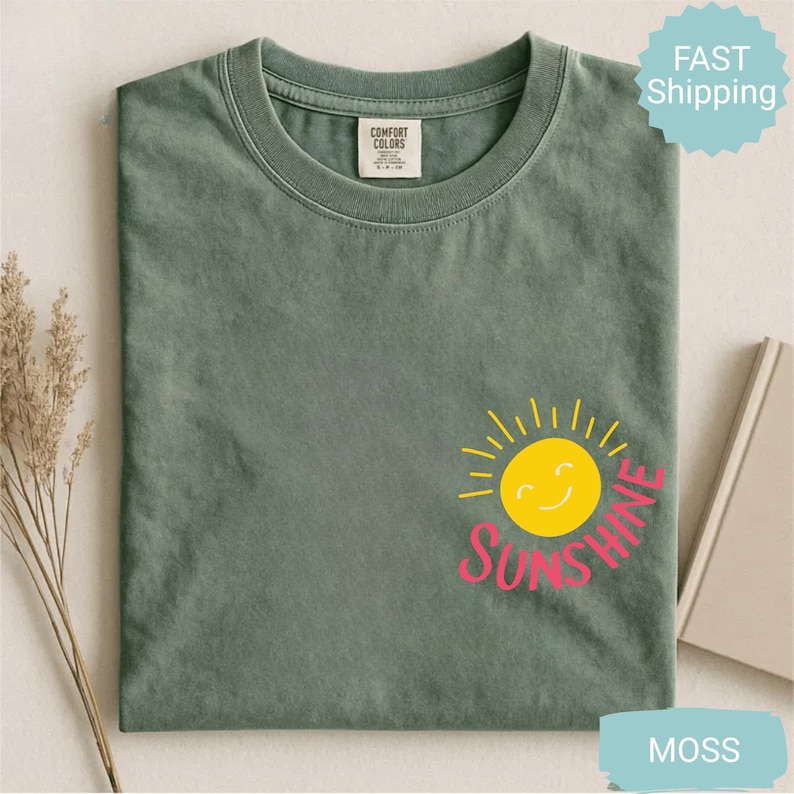 May include: A moss green t-shirt with a crew neck and short sleeves. The shirt features a smiling yellow sun graphic with the word "Sunshine" in pink. The shirt is folded and displayed on a white surface.