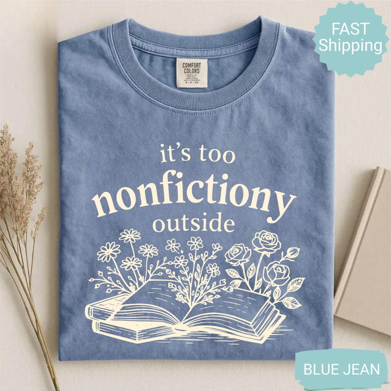 Op de afbeelding: Blauw spijkerbroekkleurig T-shirt met de witte tekst "it's too nonfictiony outside". Het ontwerp bevat een open boek met bloemenillustraties. Het shirt heeft een ronde hals en een Comfort Colors-label.