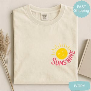 May include: Ivory-coloured t-shirt featuring a yellow smiling sun graphic and the word "Sunshine" in pink. The shirt has a crew neck and short sleeves. The shirt is made of a soft, comfortable material.