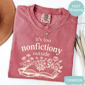 Op de afbeelding: Karmizijnrood T-shirt met de tekst "it's too nonfictiony outside" en een illustratie van een open boek met bloemen. Het shirt heeft een ronde hals en korte mouwen. Een zilveren ketting met een hanger is ook zichtbaar.