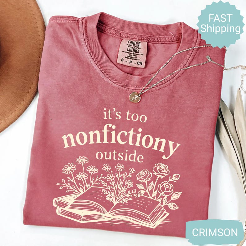 Op de afbeelding: Karmizijnrood T-shirt met de tekst "it's too nonfictiony outside" en een illustratie van een open boek met bloemen. Het shirt heeft een ronde hals en korte mouwen. Een zilveren ketting met een hanger is ook zichtbaar.