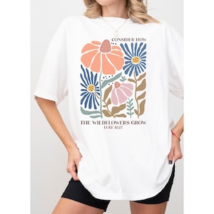 K&ouml;nnte beinhalten: Wei&szlig;es T-Shirt mit einem Blumenmuster und dem Text "Betrachte, wie die Wildblumen wachsen Lukas 12,27".