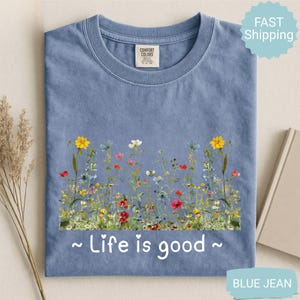 May include: A blue jean coloured t-shirt with a floral design across the front. The design features colourful wildflowers and the phrase "Life is good" in white script. The shirt has a crew neck and a Comfort Colors tag.