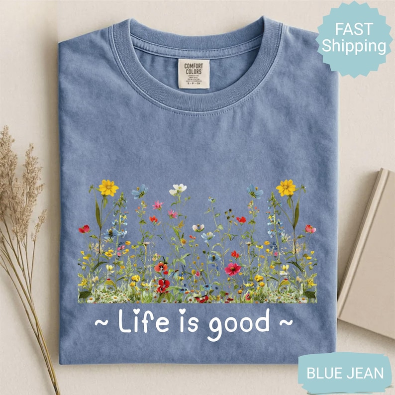 May include: A blue jean coloured t-shirt with a floral design across the front. The design features colourful wildflowers and the phrase "Life is good" in white script. The shirt has a crew neck and a Comfort Colors tag.