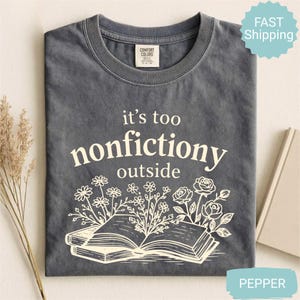 Op de afbeelding: Grijs T-shirt met de witte tekst "it's too nonfictiony outside". Het ontwerp toont een open boek en bloemen. Merk Comfort Colors.