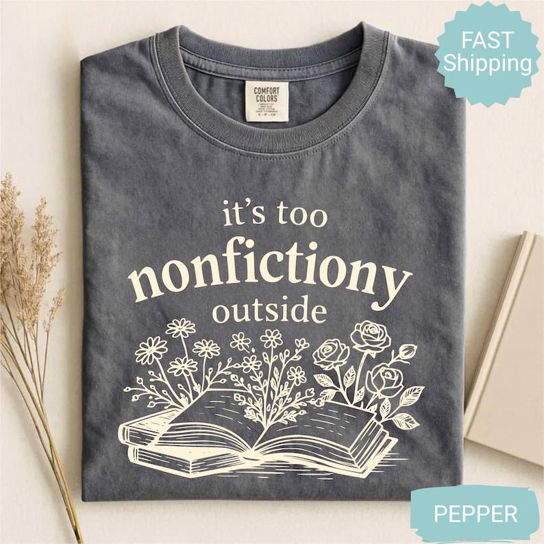 Op de afbeelding: Grijs T-shirt met de witte tekst "it's too nonfictiony outside". Het ontwerp toont een open boek en bloemen. Merk Comfort Colors.