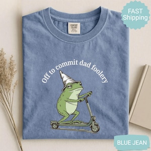 May include: Blue jean Comfort Colors t-shirt featuring a green frog in a wizard hat on a scooter. The text "Off to commit dad foolery" is arched above the frog. The t-shirt is a blue colour.