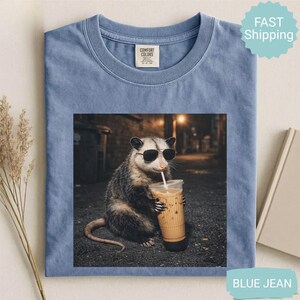 May include: Blue jean Comfort Colors t-shirt featuring a graphic of an opossum wearing sunglasses and drinking iced coffee. The opossum is in an alleyway setting. The shirt has a crew neck and a small tag on the upper left.
