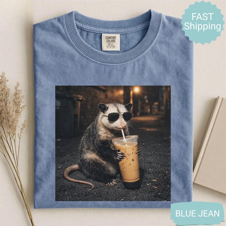 May include: Blue jean Comfort Colors t-shirt featuring a graphic of an opossum wearing sunglasses and drinking iced coffee. The opossum is in an alleyway setting. The shirt has a crew neck and a small tag on the upper left.