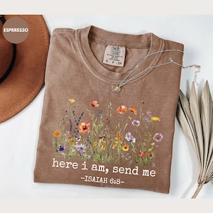 Puede incluir: Camiseta marrón con un diseño floral y el texto "here i am, send me -ISAIAH 6:8-". La camiseta tiene cuello redondo y mangas cortas. Un collar plateado con un colgante está sobre la camiseta.