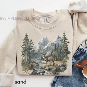 Puede incluir: Sudadera color arena con un dise&ntilde;o de monta&ntilde;a y bosque, con un ciervo cerca de un r&iacute;o. El texto "BESIMPLEBEBOLDGIFTS" est&aacute; impreso en la parte superior izquierda. La palabra "sand" est&aacute; impresa en la parte inferior.