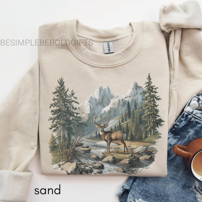 Puede incluir: Sudadera color arena con un dise&ntilde;o de monta&ntilde;a y bosque, con un ciervo cerca de un r&iacute;o. El texto "BESIMPLEBEBOLDGIFTS" est&aacute; impreso en la parte superior izquierda. La palabra "sand" est&aacute; impresa en la parte inferior.