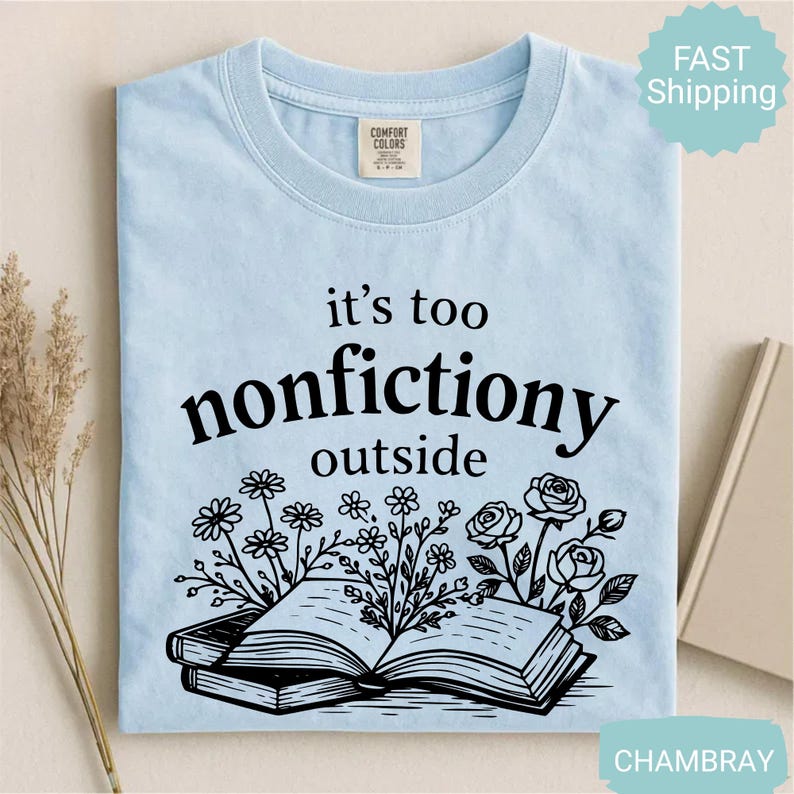 Op de afbeelding: Lichtblauw T-shirt met de zwarte tekst "it's too nonfictiony outside". Daaronder een zwarte illustratie van een open boek met bloemen en rozen. Het shirt is gemaakt van Comfort Colors materiaal.