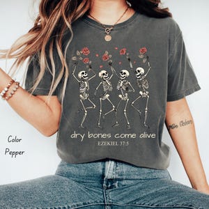 Puede incluir: Camiseta color pimienta con un gráfico de cuatro esqueletos bailando sosteniendo rosas rojas. El texto "dry bones come alive" y "EZEKIEL 37:5" están impresos debajo. La camiseta es de corte holgado.