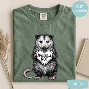 May include: Moss green t-shirt featuring a detailed illustration of an opossum holding a heart-shaped sign that reads "Absolutely Not". The opossum is depicted in black, white, and pink tones. The shirt is made of a soft material.