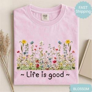 May include: A pale pink t-shirt featuring a vibrant floral design with yellow, blue, red, and white flowers. The text "~ Life is good ~" is printed in black beneath the flowers. A Comfort Colors label is visible.