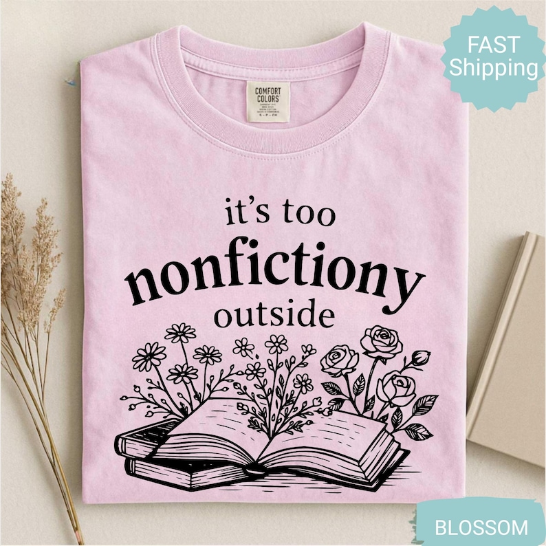 Op de afbeelding: Lichtroze t-shirt met de zwarte tekst "it's too nonfictiony outside" en een afbeelding van een open boek met bloemen. Het t-shirt is opgevouwen.