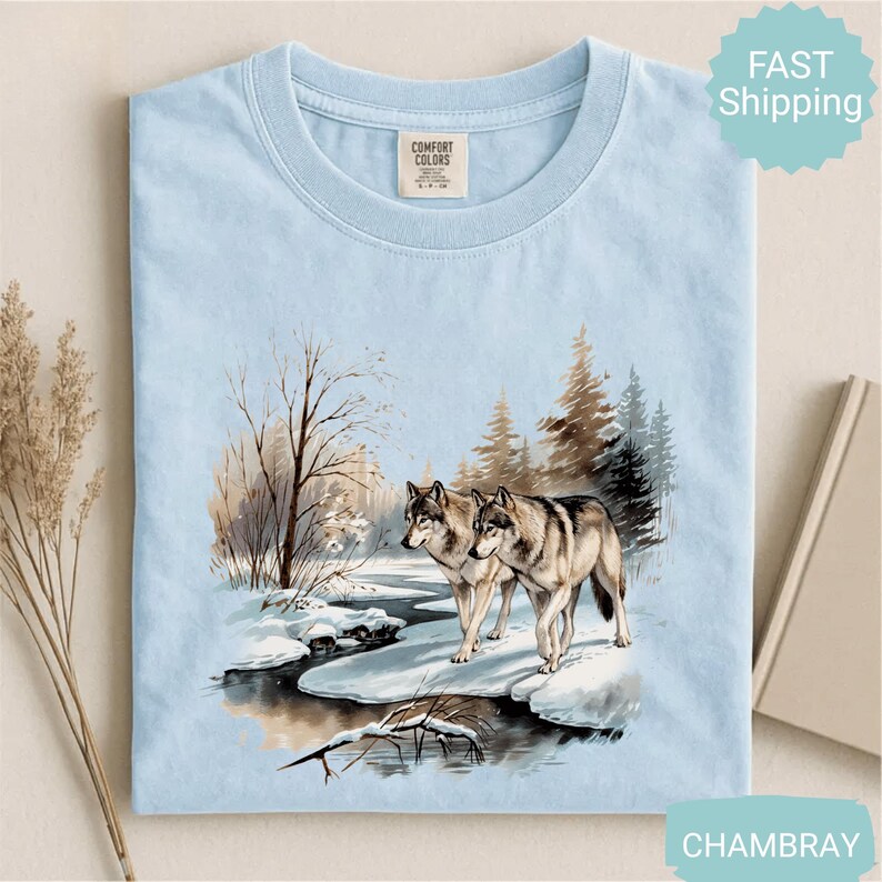 Op de afbeelding: Lichtblauw T-shirt met een winterse sc&egrave;ne met twee wolven bij een besneeuwde rivier. De afbeelding bevat bomen en een besneeuwd landschap. Het shirt heeft een ronde hals en een label "Comfort Colors".