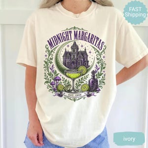 May include: An ivory t-shirt featuring a graphic print with the text "MIDNIGHT MARGARITAS" and "EST. 1998". The design includes a purple mansion, a crescent moon, a margarita glass, a lime slice, a small frog, mushrooms, and a glass bottle.