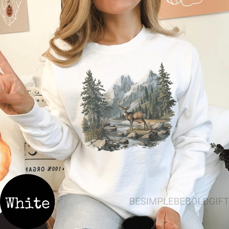 Puede incluir: Sudadera blanca de manga larga con una escena de la naturaleza que representa un ciervo, &aacute;rboles y monta&ntilde;as. La palabra "White" est&aacute; en un c&iacute;rculo negro en la parte inferior izquierda. El texto "BESIMPLEBEBOLDGIFT" est&aacute; en la parte inferior derecha.
