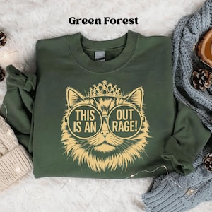 May include: Green forest-colored sweatshirt with a gold cat graphic wearing sunglasses and a crown. The graphic reads "THIS IS AN OUTRAGE!". The sweatshirt is folded on a soft, textured surface.