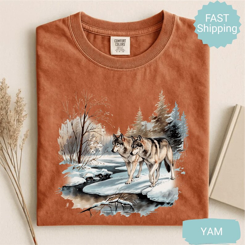 Op de afbeelding: Roestkleurig Comfort Colors T-shirt met een winterse sc&egrave;ne van twee wolven bij een besneeuwde rivier. De afbeelding bevat bomen en een besneeuwd landschap. Het shirt heeft een ronde hals en korte mouwen. De tekst "FAST Shipping" staat rechtsboven.