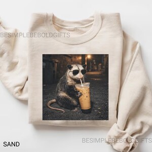 May include: Sand-colored sweatshirt featuring a graphic of an opossum wearing sunglasses and drinking an iced coffee. The image is set against a blurred street scene. The text "BESIMPLEBEOLDGIFTS" is printed on the sweatshirt.