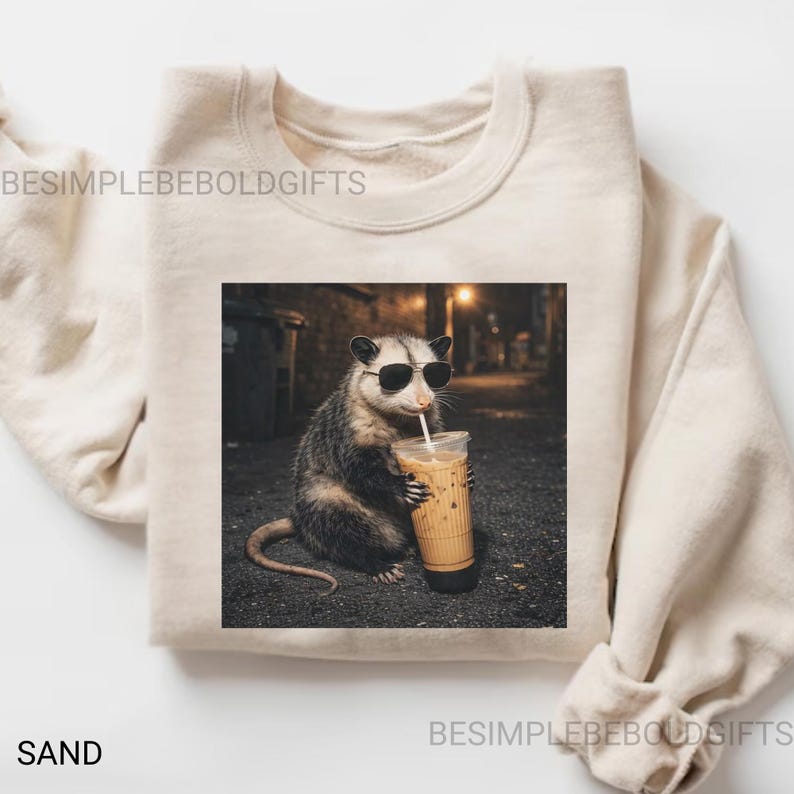 May include: Sand-colored sweatshirt featuring a graphic of an opossum wearing sunglasses and drinking an iced coffee. The image is set against a blurred street scene. The text "BESIMPLEBEOLDGIFTS" is printed on the sweatshirt.
