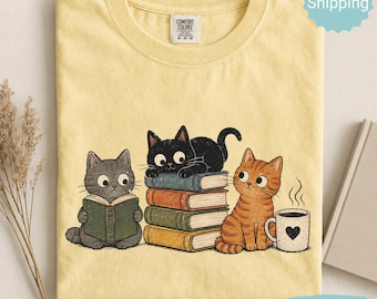 May include: A pale yellow t-shirt featuring a graphic of three cats. One gray cat reads a green book, a black cat rests on a stack of four colorful books, and an orange tabby cat sits beside a white mug with a black heart and steam rising.