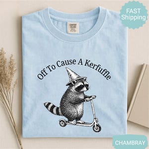 May include: Light blue t-shirt with a graphic of a raccoon wearing a witch hat and riding a scooter. The text "Off To Cause A Kerfuffle" is arched above the raccoon. The shirt has a crew neck and short sleeves.