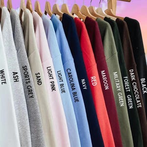 May include: A selection of sweatshirts in a variety of colours, displayed on wooden hangers. The colours include white, ash, sports grey, sand, light pink, light blue, Carolina blue, navy, red, maroon, military green, forest green, dark chocolate, and black.