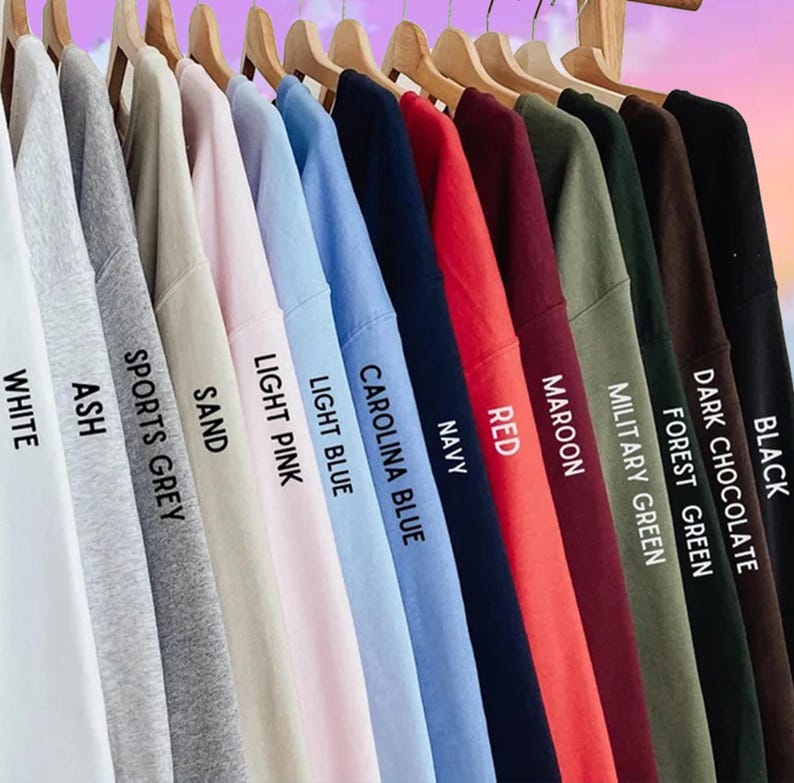 May include: A selection of sweatshirts in a variety of colours, displayed on wooden hangers. The colours include white, ash, sports grey, sand, light pink, light blue, Carolina blue, navy, red, maroon, military green, forest green, dark chocolate, and black.