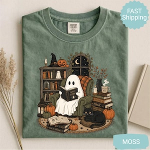 May include: A sage green t-shirt featuring a graphic illustration of a white ghost sitting in an armchair reading a book titled Ghost Stories. The scene includes a bookshelf, a black cat, pumpkins, candles, and a stack of books on a round rug.