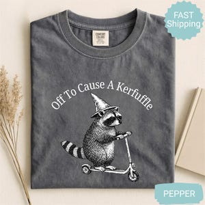 May include: A dark gray t-shirt with the text "Off To Cause A Kerfuffle" above a raccoon riding a scooter and wearing a wizard hat. The shirt is made of a soft material.