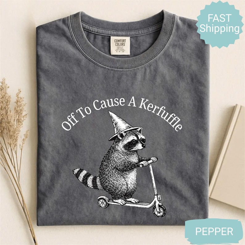 May include: A dark gray t-shirt with the text "Off To Cause A Kerfuffle" above a raccoon riding a scooter and wearing a wizard hat. The shirt is made of a soft material.