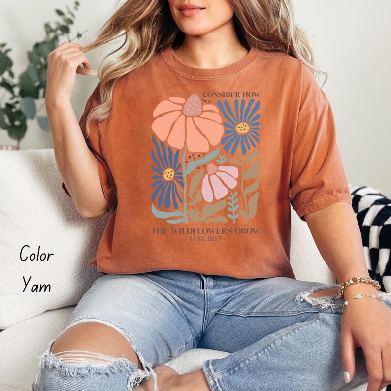 K&ouml;nnte beinhalten: Ein rostfarbenes T-Shirt mit einem Blumendesign und dem Text "Consider how the wildflowers grow" in Wei&szlig;.