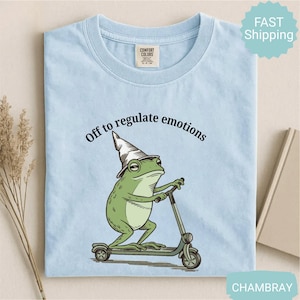 May include: Pale blue t-shirt featuring a green frog in a wizard's hat on a scooter, with the text "Off to regulate emotions". The shirt is made of a soft material. The shirt is a light blue colour.