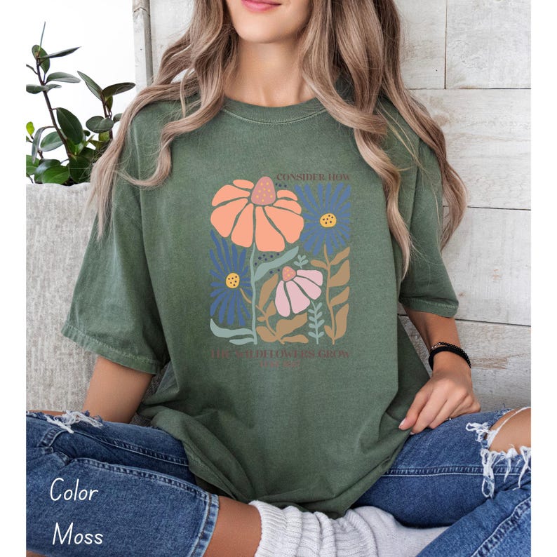 K&ouml;nnte beinhalten: Ein moosgr&uuml;nes T-Shirt mit einem floralen Motiv und dem Text "Consider how the seed of flowers grow" auf der Vorderseite.