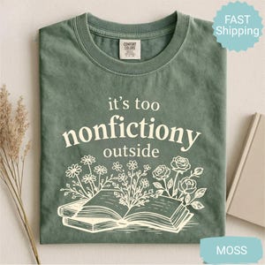 Op de afbeelding: Mosgroen t-shirt met de witte tekst "it's too nonfictiony outside". Het ontwerp bevat een open boek met bloemenillustraties. Het shirt heeft een ronde hals en een label op de bovenrug.