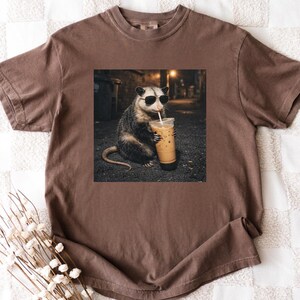 May include: Brown t-shirt featuring a graphic of an opossum wearing sunglasses and drinking iced coffee. The opossum is in a dark alley setting. The shirt is made of a soft material.