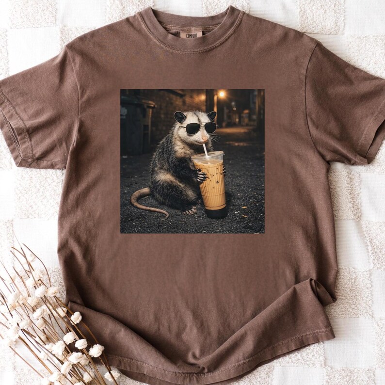 May include: Brown t-shirt featuring a graphic of an opossum wearing sunglasses and drinking iced coffee. The opossum is in a dark alley setting. The shirt is made of a soft material.