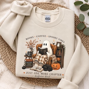 May include: A sand-colored crewneck sweatshirt featuring a graphic of a ghost sitting in an armchair reading a book. The design includes text that reads BOOKS COFFEE SPOOKY VIBES and JUST ONE MORE CHAPTER before bed, along with pumpkins and books.